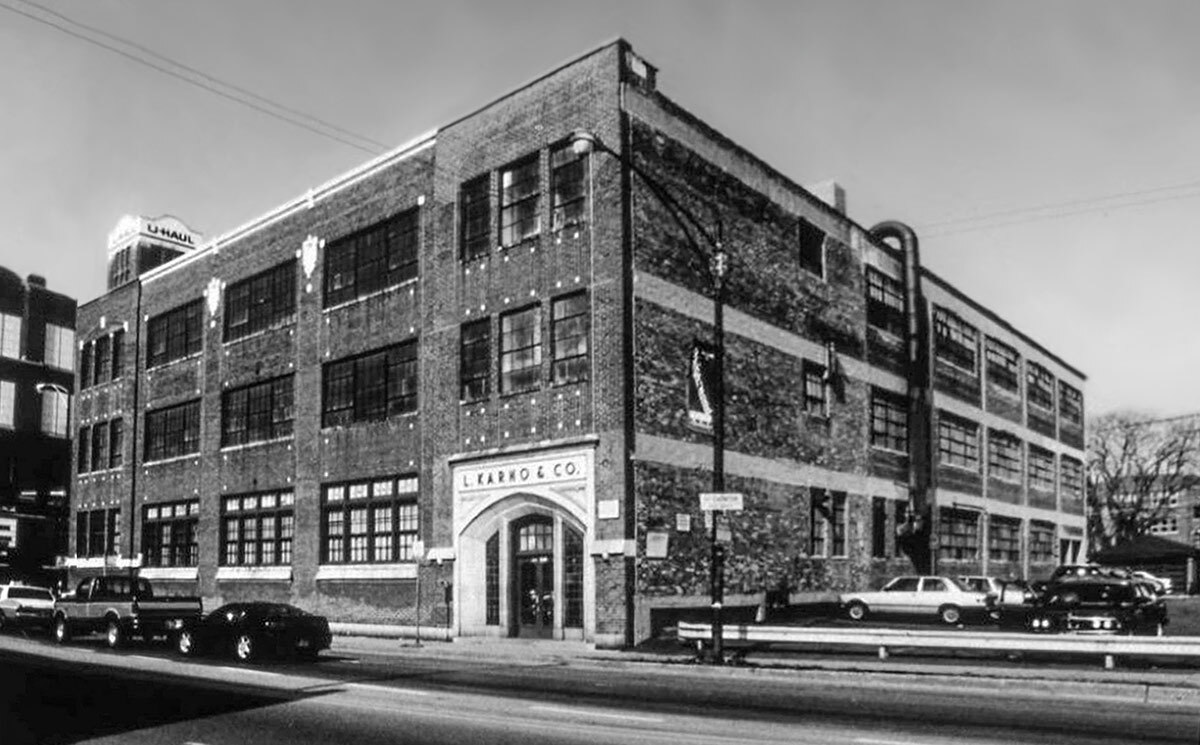 The Fullerton Building on the Lincoln Park Campus is shown in 1992. The building is on a street corner lined with old cars and a street lamp. It is made of bricks and has three floors of rectangular windows with square panels. The front door is on the left side of the building and framed by a concrete archway. Above the door is a sign that reads “L. Karno & Co.”