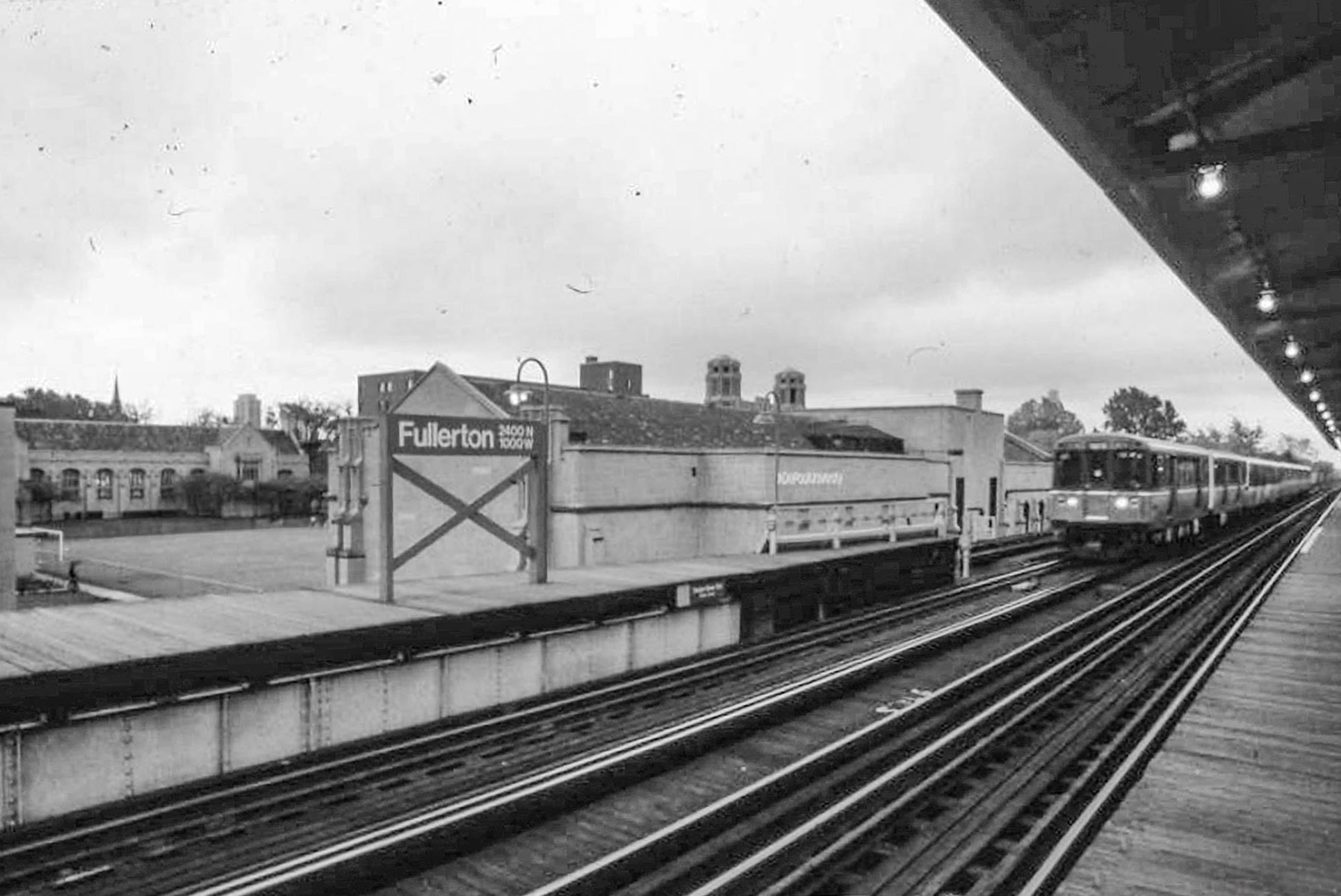 The Fullerton Station on DePaul’s Lincoln Park Campus is shown in 1978. The station has wooden platforms on either side of the tracks. A CTA train is approaching the station. Behind the station is an athletic field, surrounded by buildings with triangular roofs.