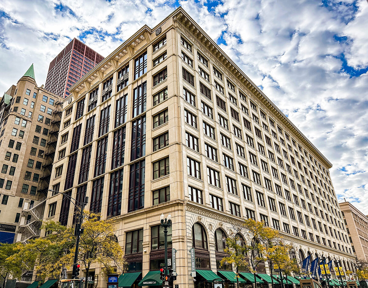 The DePaul Center is shown in 2022. The building is on a street corner surrounded by other skyscrapers and trees with yellow leaves. The beige-brown stone clad building has green awnings over the first floor windows. The former CNA Building, known informally as “Big Red,” is shown peeking over the top of the DePaul Center.
