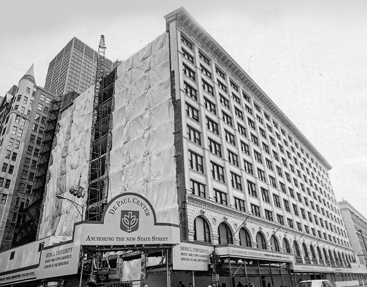 The DePaul Center in Chicago’s Loop neighborhood is shown under construction in 1992. The building has 11 floors of rectangular windows and arched windows on the lowest floor. One side of the building is covered in a tarp and scaffolding. A sign in front on the building reads, “DePaul Center, Anchoring the New State Street.”